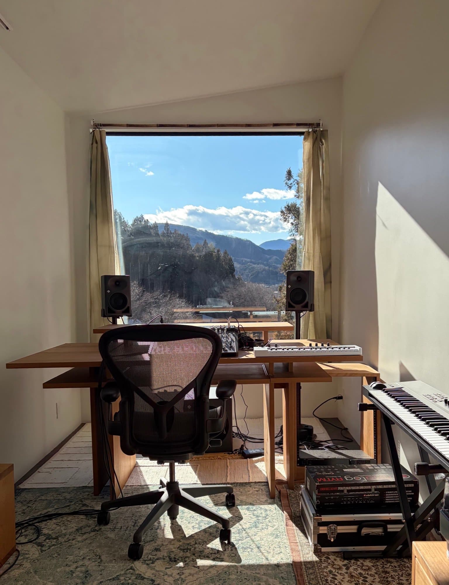 산줄기가 보이는 큰 창 앞에 놓인 컨트롤룸 데스크. Neumann 모니터와 MIDI 키보드
